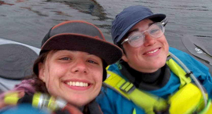 Two people wearing life jackets sit in boats and smile for the photo. 